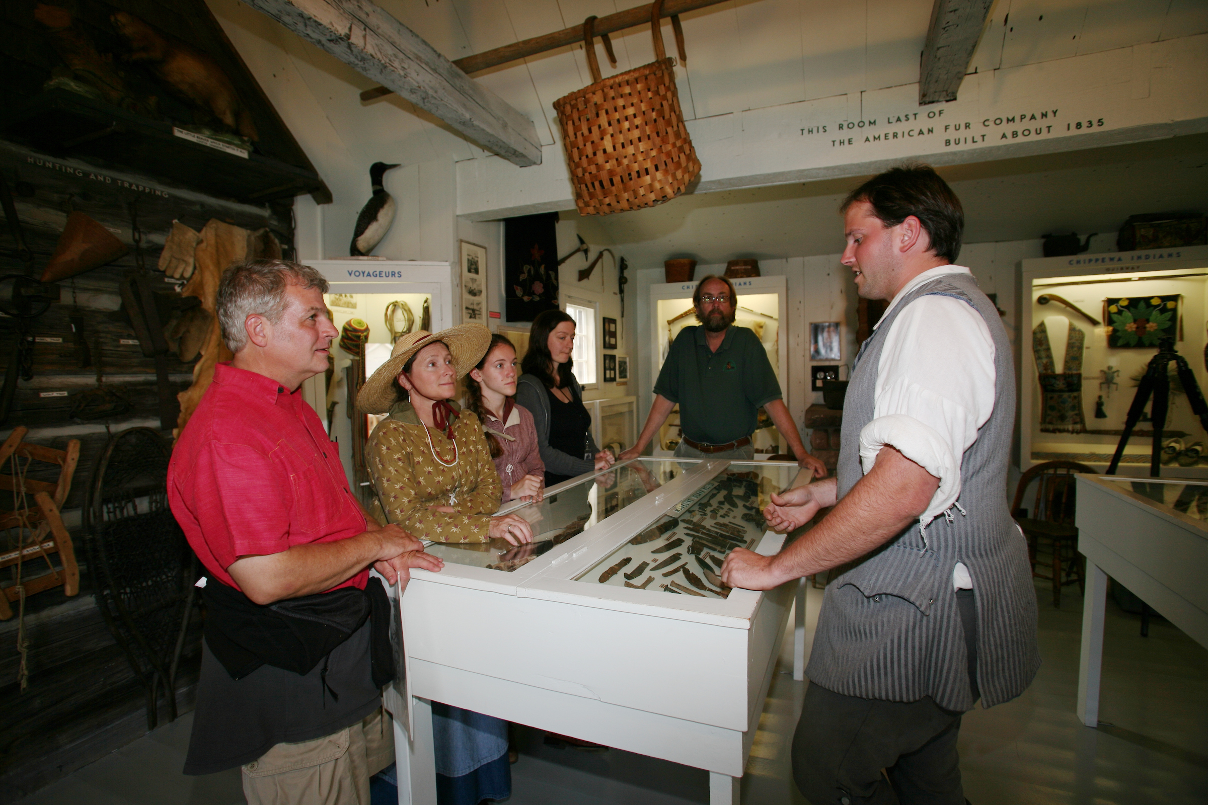 A group of white people stand around a display case with a museum volunteer describing the contents.